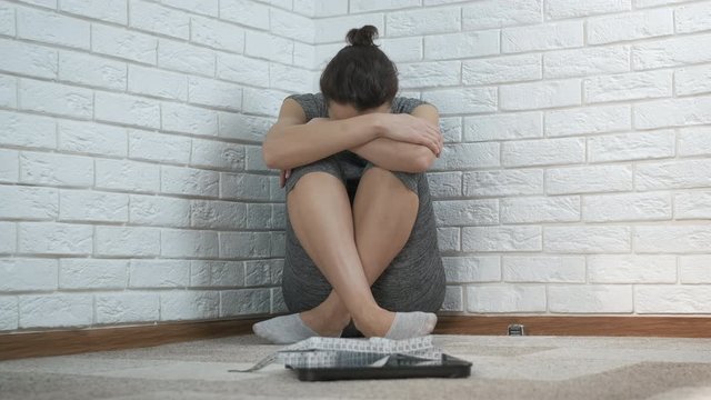 Overweight Depression. A depressed woman sits in the corner of a room in front of a scale.