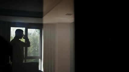 One young man drinks whiskey alcohol while standing by a large window at home. Shooting in backlight through the glass. - Powered by Adobe