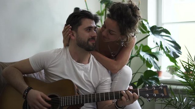 Young woman and man playing guitar while sitting on sofa in modern apartment spbd. Beautiful American couple has good time together, pretty female kisses bearded guy tenderly on couch in white