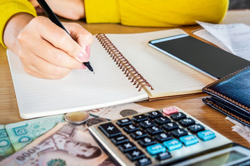 woman hand holding pen writing on notebook with money, calculator and smart phone on desk...