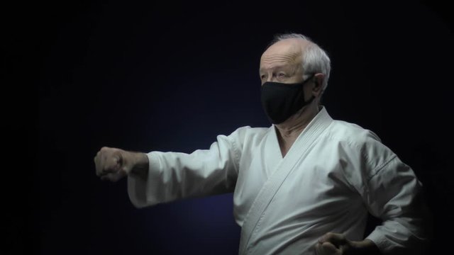 In a black medical mask, an old athlete is training blocks and punches against a dark background