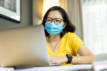 Asian female wear eyeglasses using laptop computer during quarantine at house. Young Woman in yellow shirt wear blue surgical mask working from home during pandemic virus. Covid 19, Coronavirus, 