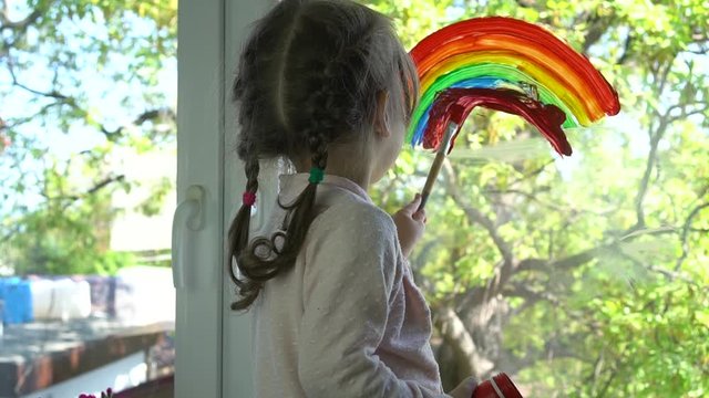 A little girl draws a rainbow on a window using paints and brush. Colourful rainbow pictures drawings on windows and balconies spread hope during coronavirus pandemic. Covid-19 outbreak. Home lockdown