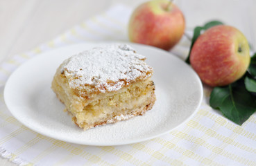 A piece of apple pie on a white plate and apples next to it. 