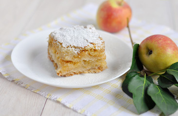 A piece of apple pie on a white plate and apples next to it. 