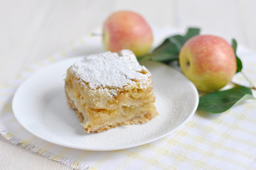 A piece of apple pie on a white plate and apples next to it. 