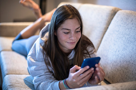 Cute Teen Girl Texting On A Smartphone Lying On A Couch At Home. Candid Indoor Photo With Focus On The Foreground And Copy Space