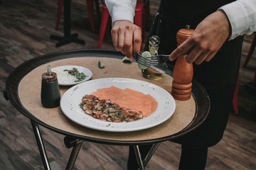 Mesero preparando carpaccio mixto de pulpo con salmón ahumado sobre bandeja. 