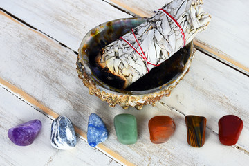 A close up image of healing chakra crystals with a white sage bundle in an abalone shell. 
