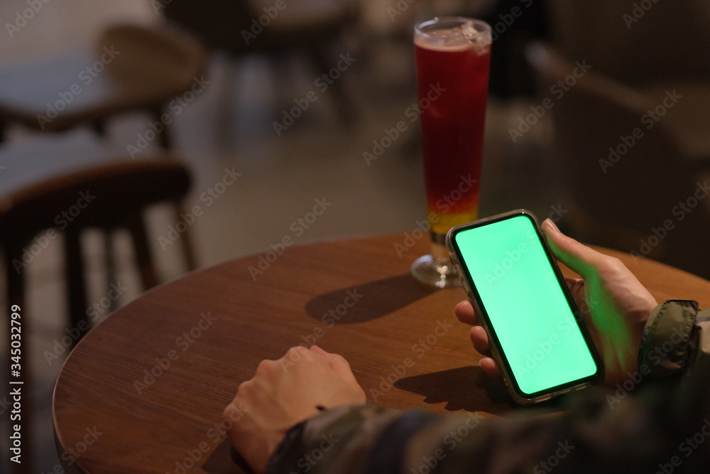 Wall mural Over shoulder of man holding green screen smart phone in the bar at night. A glass of cocktail on woode table. Blur background