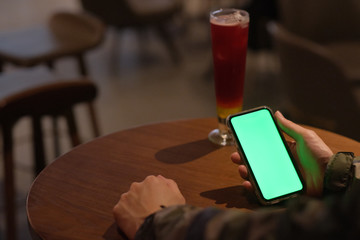 Over shoulder of man holding green screen smart phone in the bar at night. A glass of cocktail on woode table. Blur background