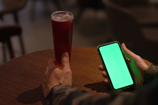 Over shoulder of man holding green screen smart phone in the bar at night. A glass of cocktail on woode table. Blur background
