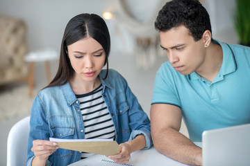 Dark-haired female and brunette male sitting at desk, checking papers together, looking concerned