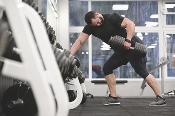 strong young bearded caucasian man in black sportswear pulling heavy weight dumbbell with one hand...
