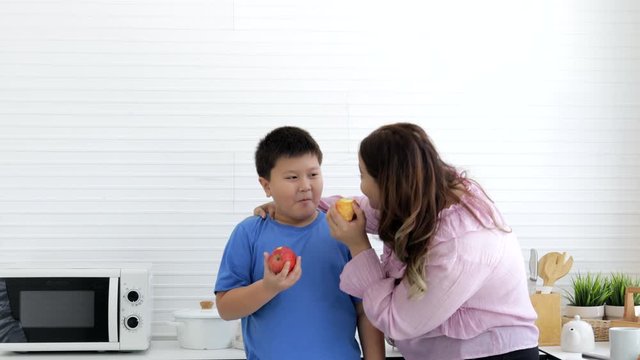 Mother and son eating ripe apple deliciously and funny together in kitchen for diet and slim body. Fat woman with healthy fruit and diet food and low calorie fruit concept