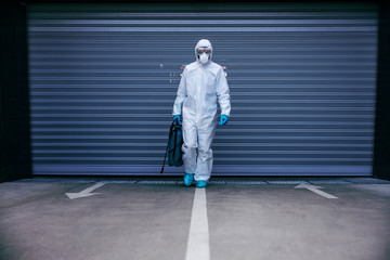 Man in sterile protective suit and mask holding disinfectant and walking towards camera in underground garage.