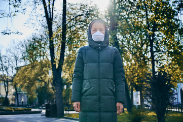 Portrait of a woman in a mask, standing outside in empty park, green trees in sunlight on a background. Social distancing during global coronavirus quarantine