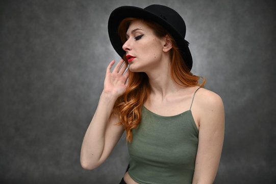 Woman shows emotions changing poses. Portrait of a young pretty girl with beautiful hair and great makeup in a green T-shirt and black hat on a gray background.