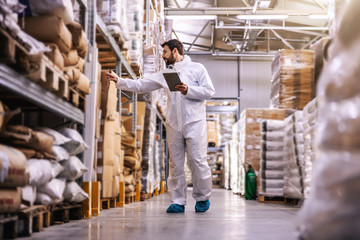 Full length of young employee in sterile protective uniform checking on products while standing in warehouse full of food products in boxes.