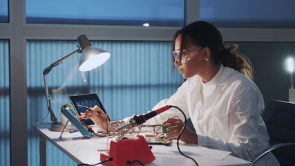 Close up of female electronics engineer in protective glasses checking motherboard with multimeter...