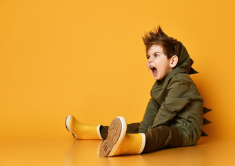 Kid boy in green hoodie with dinosaurs spikes at his back and cowl sits on the floor roaring loud...
