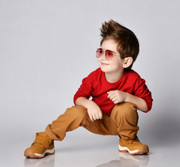 Smiling little kid boy in red jumper, sunglasses, brown pants and sneakers sits squatting looking...