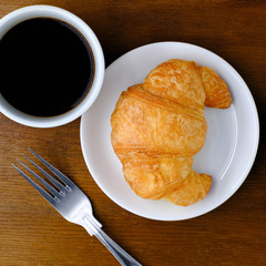 The cup of black coffee and delicious french homemade Croissant on a white plate with fork on wooden background in the morning sunlight, Top view with copy space, Yummy and healthy concept.