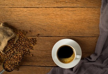 Top view above of Black hot coffee for morning with milk foam for morning menu in white ceramic cup with coffee beans roasted in burlap sack bag on wood table background. Flat lay with copy space.