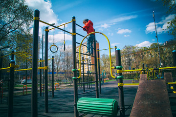Fototapeta na wymiar A boy in a red jacket is engaged in a sports field in the Park in the spring, climbing up the stairs. Swedish wall. Trips, sports classes for children, physical education, games in the fresh air.