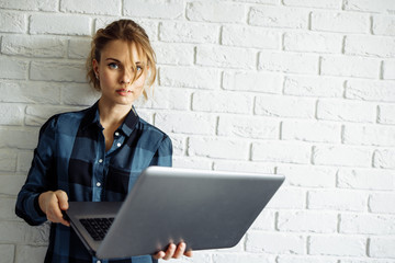 Young woman freelancer with laptop in her hands standing against white brick wall.