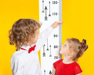 The older brother measures the height of the younger sister using a height meter. Slavic, European children on a yellow background. A boy in a red bow tie. A girl in a red dress.