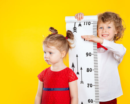 The older brother measures the height of the younger sister using a height meter. The girl is dissatisfied with her height. My brother is laughing and happy. Slavic, European children.