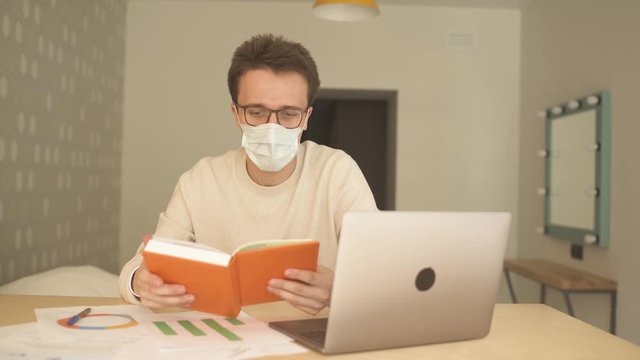 Young man in medical mask, glasses and white sweater doing a research with his laptop and writing notes in his notebook. People have to work and study  from home during covid-19 coronavirus