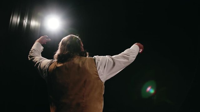 Rear view of an adult male actor with long hair playing a role on stage in front of a dark auditorium against the background of spotlights. Expressive emotion and hand gestures. Inside the theater