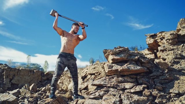 Strong man is training with heavy hammer. Athletic sportsman breaking stones with iron hammer on the natural rocky background. Slow motion.