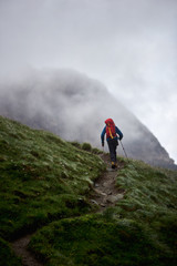 Back view of man backpacker with trekking sticks climbing the grassy hill. Traveler with backpack walking on path and heading to foggy mountain. Concept of hiking, climbing and backpacking.