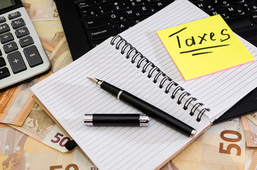 inscription taxes on a sticker, notebook, pen and part of a laptop on a background of euro banknotes. Business concept.