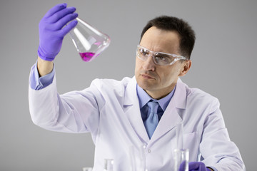 40s Male doctor or Chemist researcher in blue gloves and protective glasses scrutinizing transparent red liquid in flask isolated on grey background chemical research, creating new materials