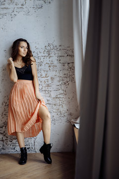A girl in spring clothes stands at the window . She leans against the loft-style wall. The girl lifts the hem of her skirt and exposes her leg. The leg is slender and beautiful. Photo for story format