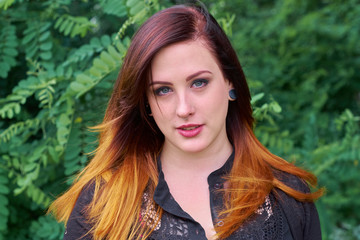 Powerful young woman with gray eyes and an ombre hairstyle coloring with tones of crimson red and orange, giving her a stunning look against green forest bush leaves.