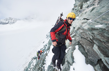 Full length of male mountaineer in sunglasses using fixed rope to climb winter mountain. Alpinist in safety helmet standing on rock covered with snow. Concept of mountaineering, alpine rock climbing.
