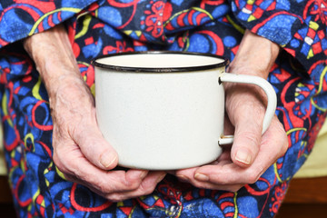 In her hands, an old grandmother of 90 years holds in her hand a metal mug with water, poverty and...