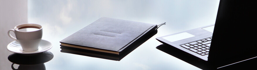 Business accessories on a glass table. A laptop and a leather-bound notebook. Long banner. Reflection of the sky. Copy of the space.