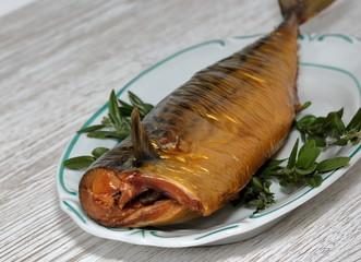 Smoked mackerel on decorative plate, few green herbst around, on wooden table close up