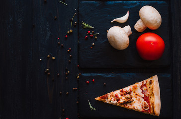 Slice of fresh homemade pizza with sun-dried tomatoes, chicken, cheese and chili peppers on a black stone plate on black wooden background. Flat lay. Place for text