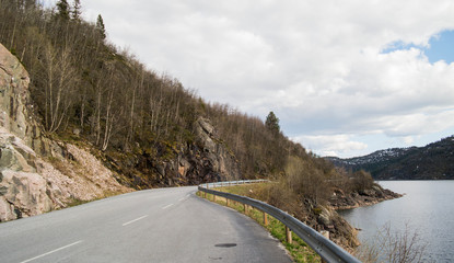 The Norwegian road along the lake and grey rocks. A beautiful spring landscape. Coniferous trees growing on the slope.