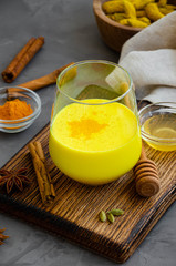 Golden milk in a glass on a wooden board with honey and other spices. A healthy drink made from milk and turmeric. Vertical orientation. Copy space.