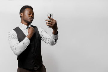 Black bearded african is looking to self through the camera on smartphone and straightens a tie. Man in white shirt and black vest on white background