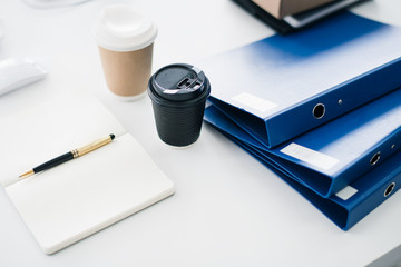Blue file folder, coffee cup, note book and pen put on the white table in home office. Business report paper file. worker deck background.