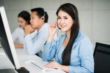 Young beautiful Asian operator woman with headsets working with team in call center.Customer support phone looking at camera with smiling and feeling happy.Team operator call center service concept.